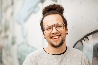 A quirky looking young man with glasses and his hair in the bun smiles at the camera, leaning against a graffiti covered wall