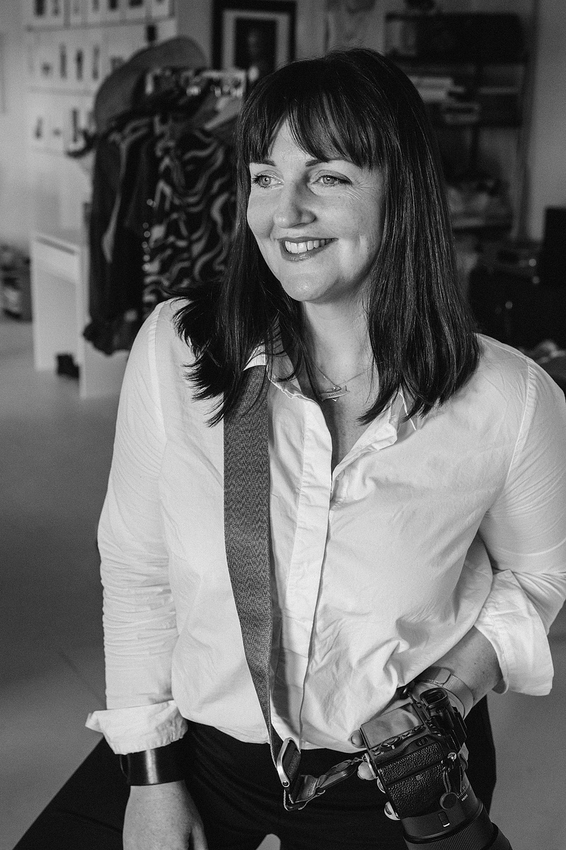 A brunette photographer smiles to the side, her camera strung across her torso