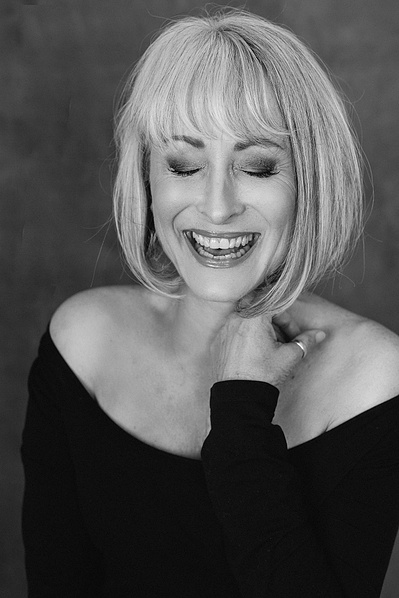 A striking blonde woman laughs, one hand resting on her collarbone