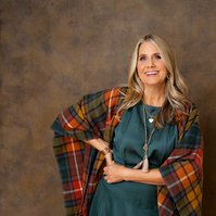 A woman with shiny blonde hair is wrapped in a tartan blanket and smiles at the camera