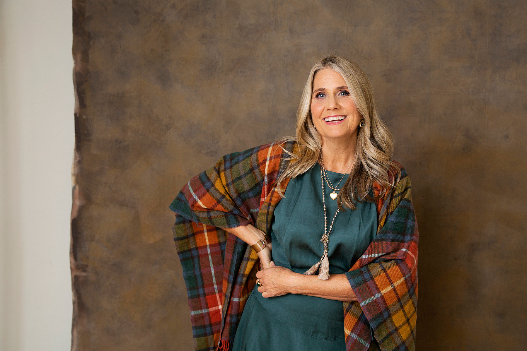 A woman with shiny blonde hair is wrapped in a tartan blanket and smiles at the camera
