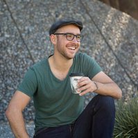 A hipster looking man sits with a cup of coffee, smiling off camera