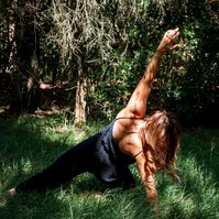 A slim woman practices yoga outside in long grass illuminated by dappled sunlight, her weight is on one knee as she stretches the other leg out and reaches an arm over her head 