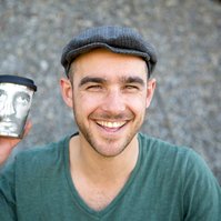 A man wearing a flat cap beams at the camera, holding a coffee cup level with his face to show that the cup is illustrated with a face