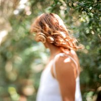A slim woman with curled hair wears a white tank top, she stand in greenery and has dappled sunlight all around her