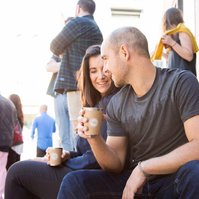 Two people sit on the kerb of a bus pedestrian street, they are enjoying a takeaway coffee together