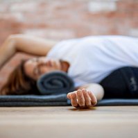A woman lies on a yoga mat using props to help her stretch, one arm is out straight towards the camera and only her hand is fully in focus