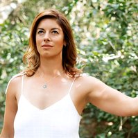 A slim woman stands in front of greenery, one arm outstretched with her index finger and thumb touching 