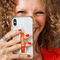 A woman with blonde curls smiles from behind her phone, it looks as though she is snapping a photo of you