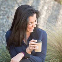 A brunette woman sits and holds a coffee in a takeaway cup, she is laughing at something