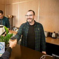 Two people work in a coffee shop, one is making coffee and the other is serving customers with a smile