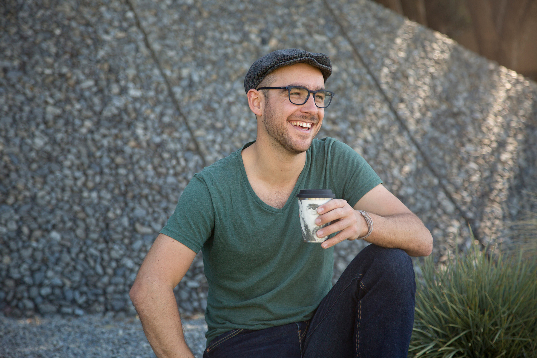 A hipster looking man sits with a cup of coffee, smiling off camera