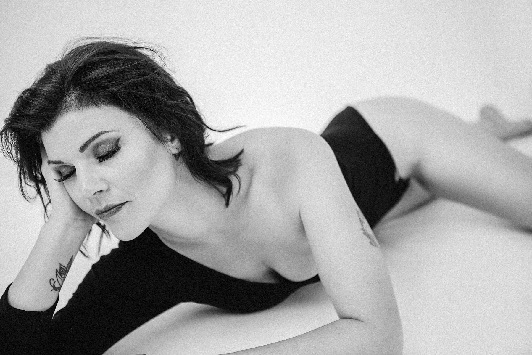 A beautiful woman with a brunette bob poses in an asymmetrical black body suit, she is laying on a bed with a hand supporting her head