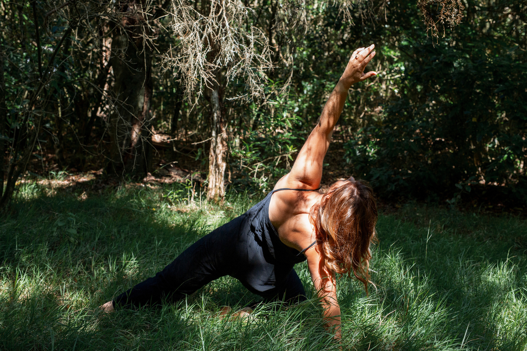 A slim woman practices yoga outside in long grass illuminated by dappled sunlight, her weight is on one knee as she stretches the other leg out and reaches an arm over her head 