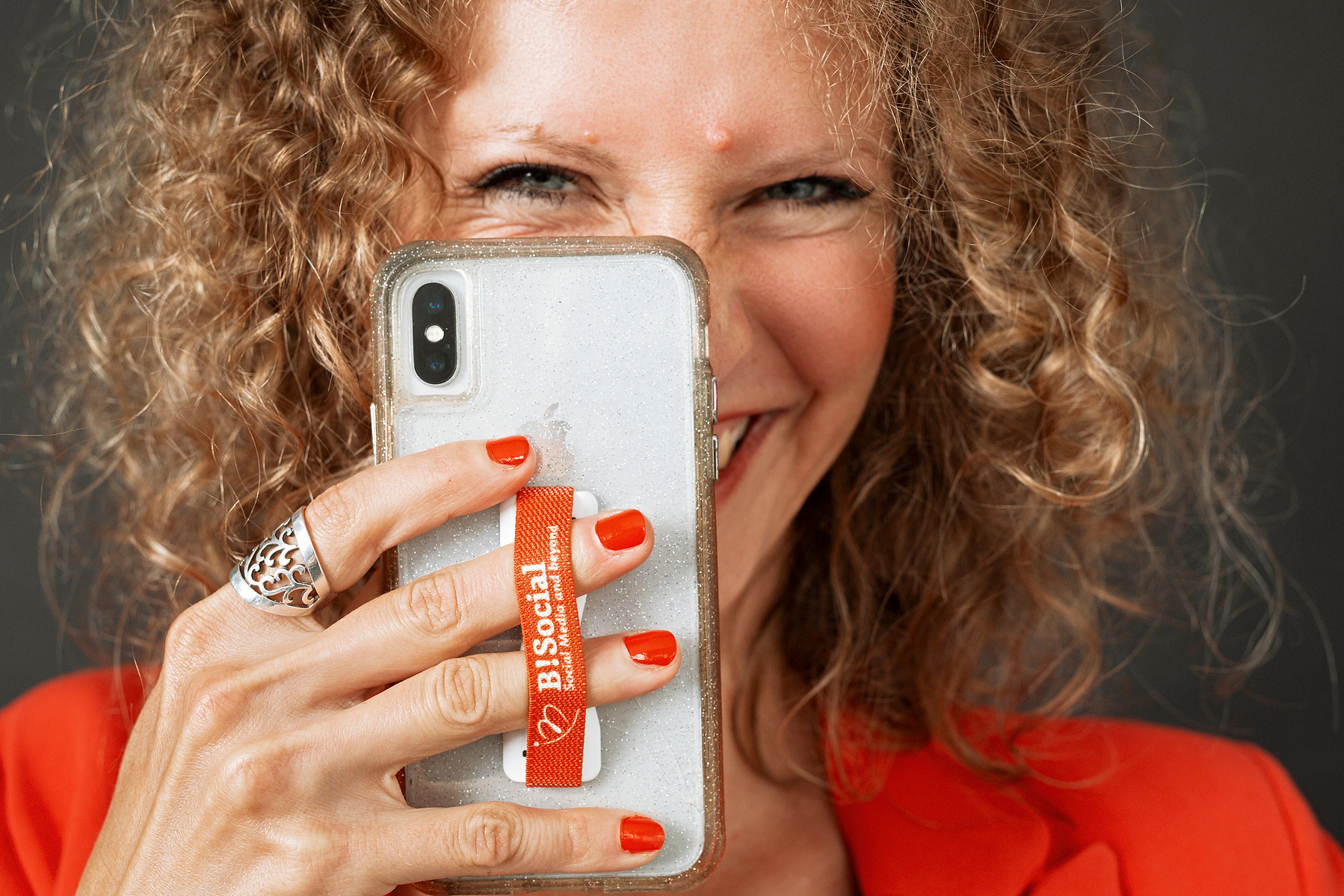 A slim woman with curly reddish hair smiles from behind her phone, as if she's taking a photo of you