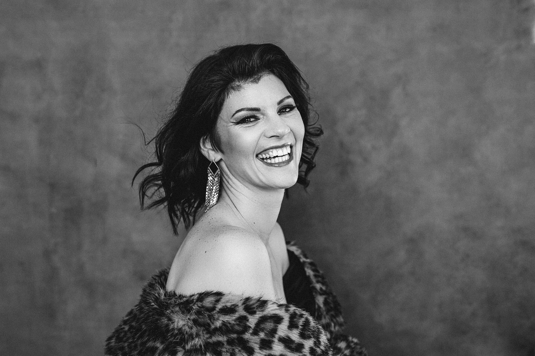 A glamourous brunette woman is wrapped in a fur with her shoulders visible, she is laughing