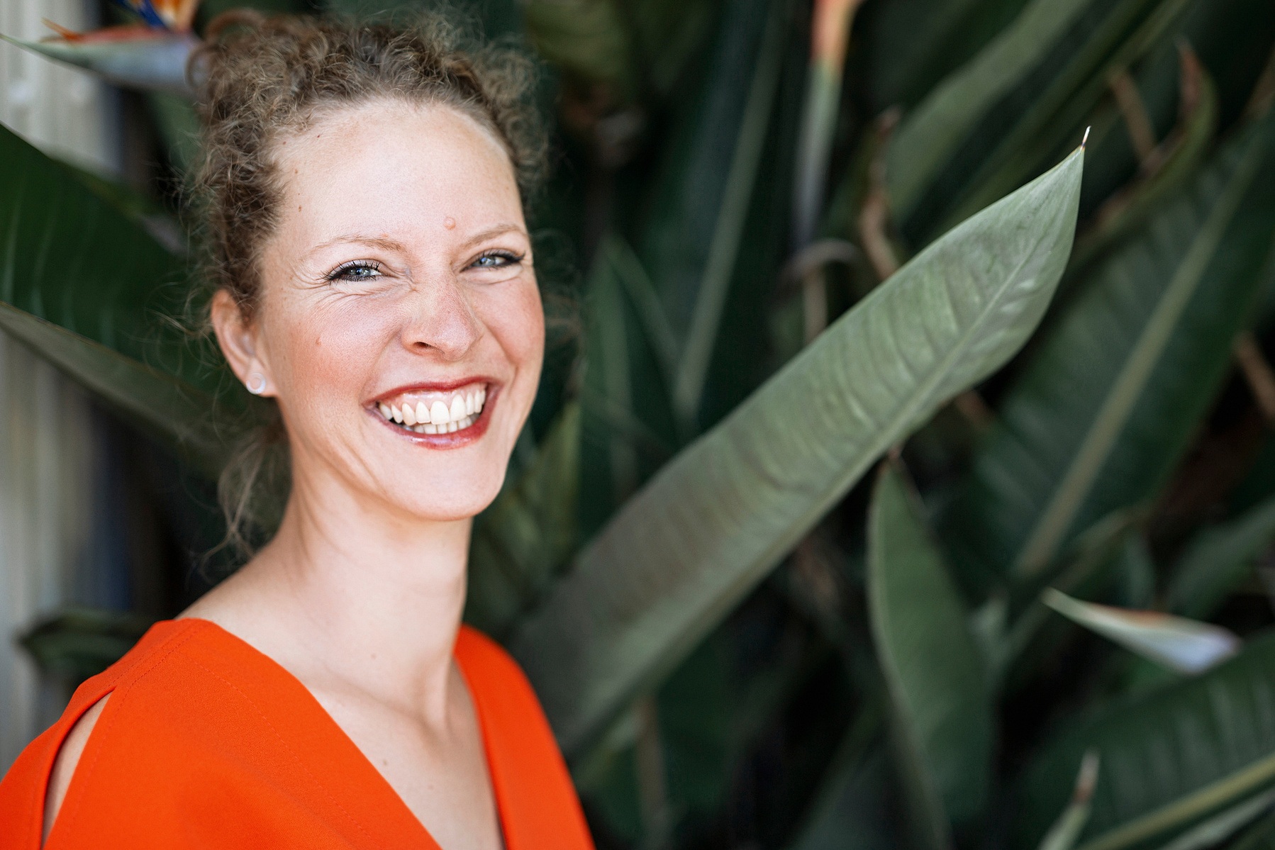 A slim woman with curly reddish hair stands in front of a tropical plant and smiles at the camera