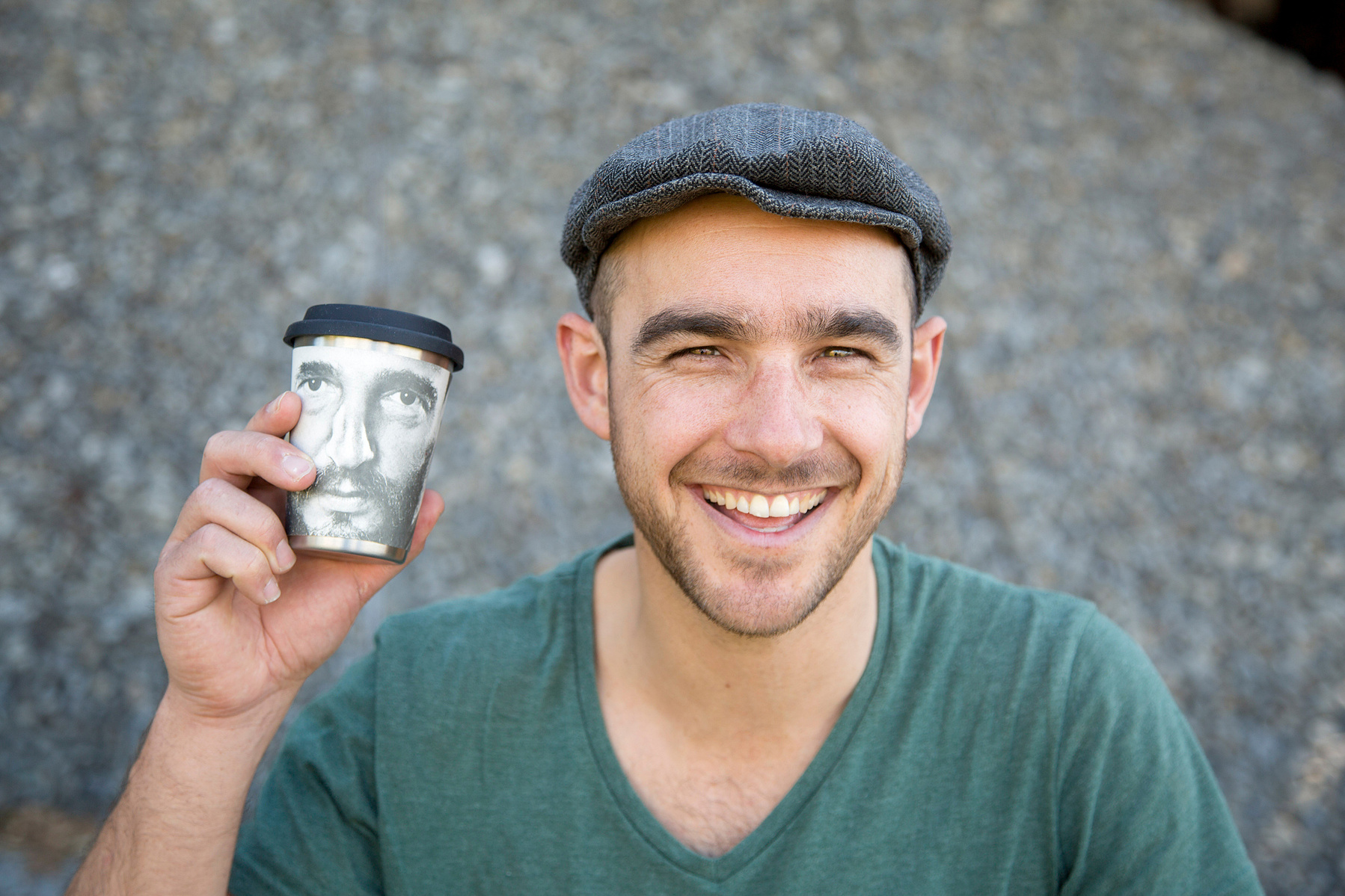 A man wearing a flat cap beams at the camera, holding a coffee cup level with his face to show that the cup is illustrated with a face
