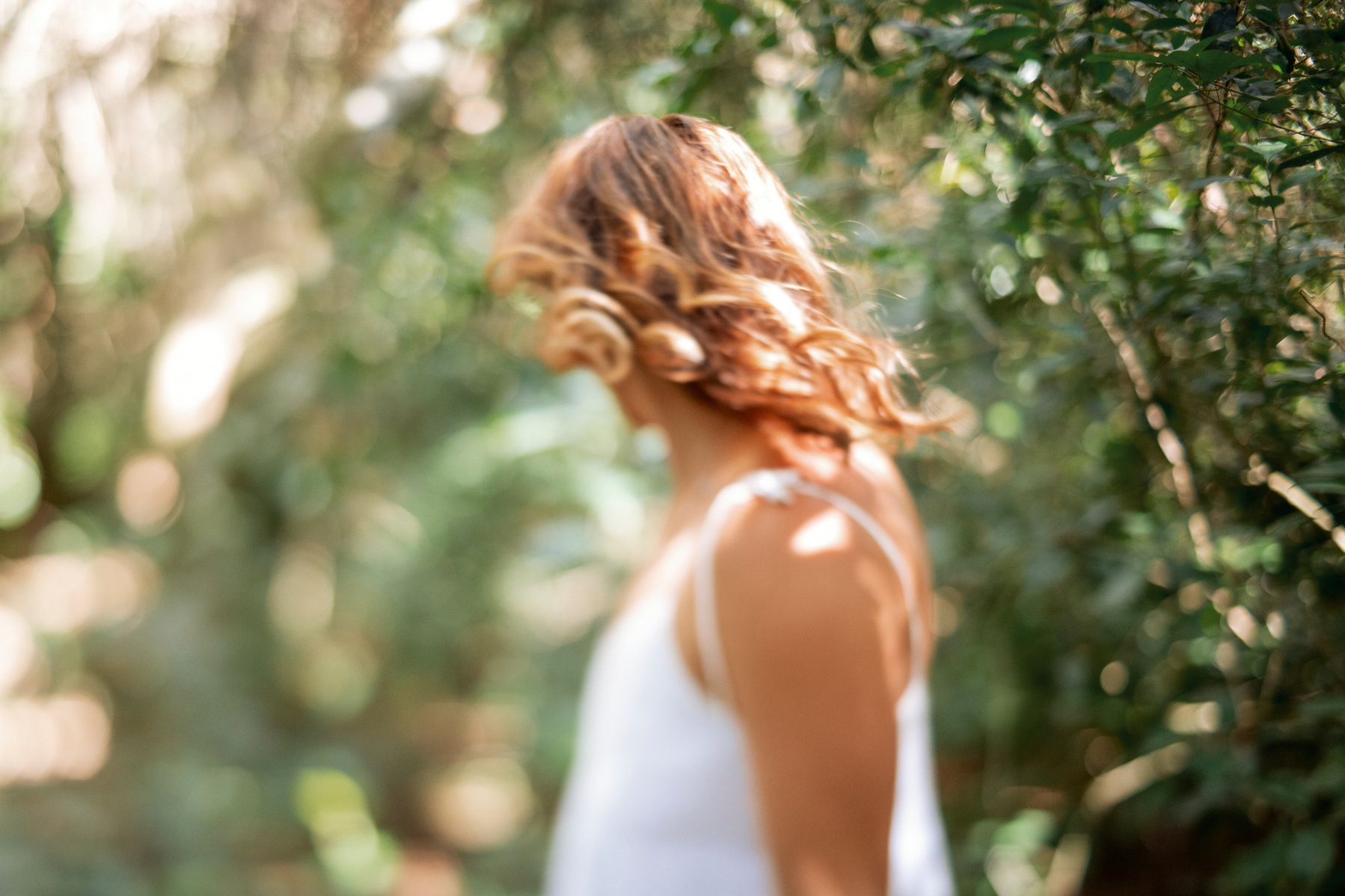 A slim woman with curled hair wears a white tank top, she stand in greenery and has dappled sunlight all around her