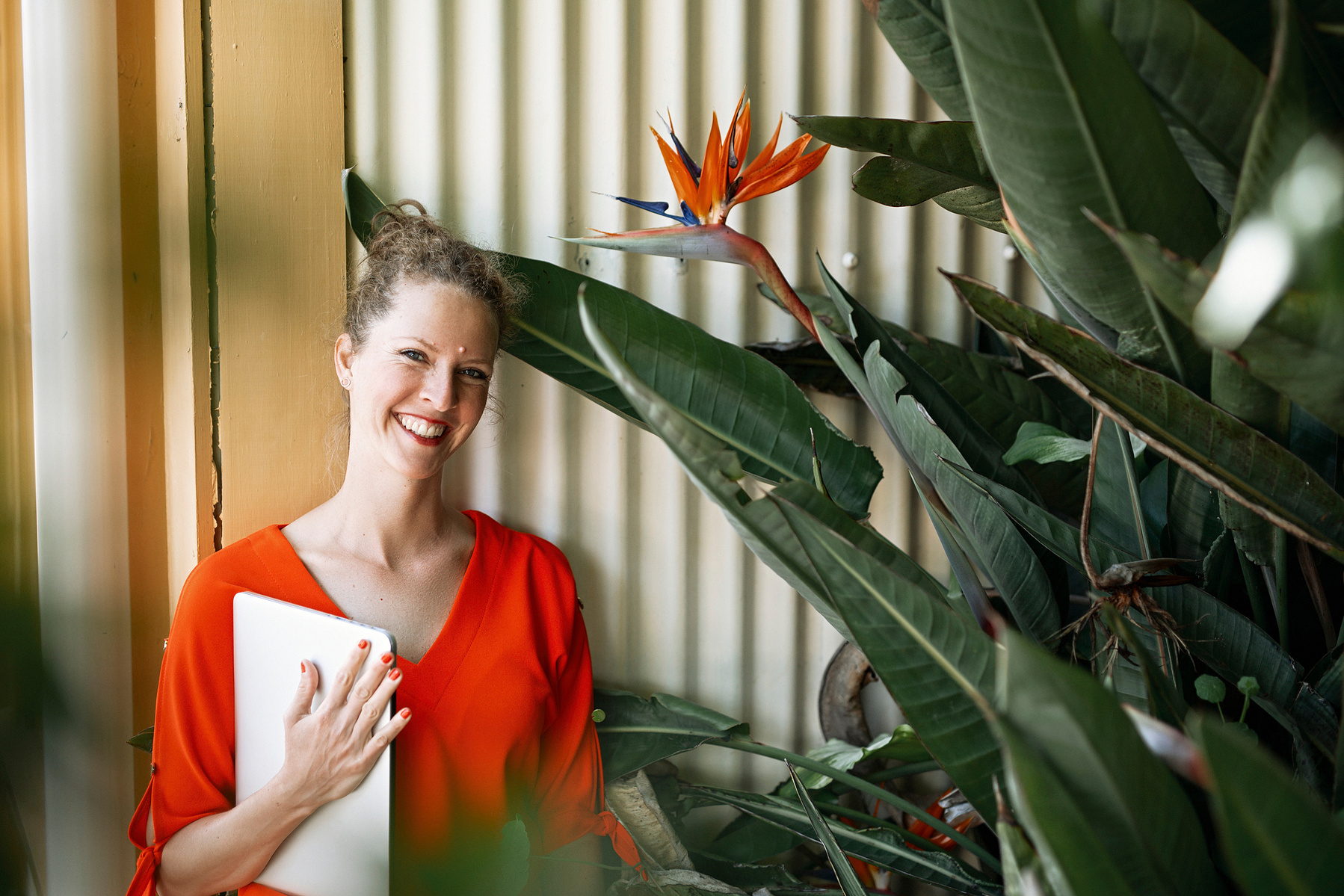 A slim woman with curly reddish hair holds a macbook and smiles behind some tropical plants