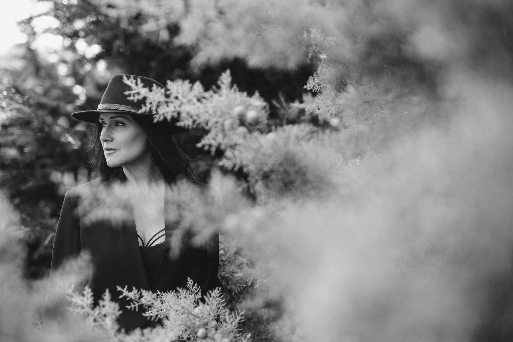 A glamourous brunette wears a black hat, she is framed by the branches of frosty pine trees