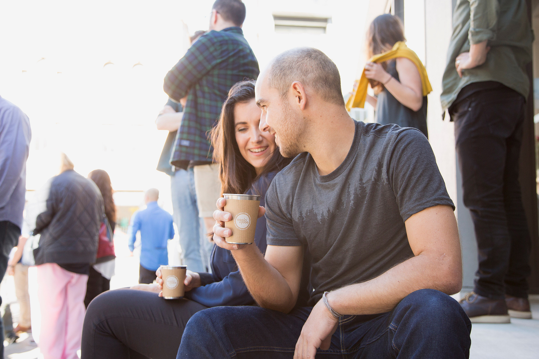 Two people sit on the kerb of a bus pedestrian street, they are enjoying a takeaway coffee together