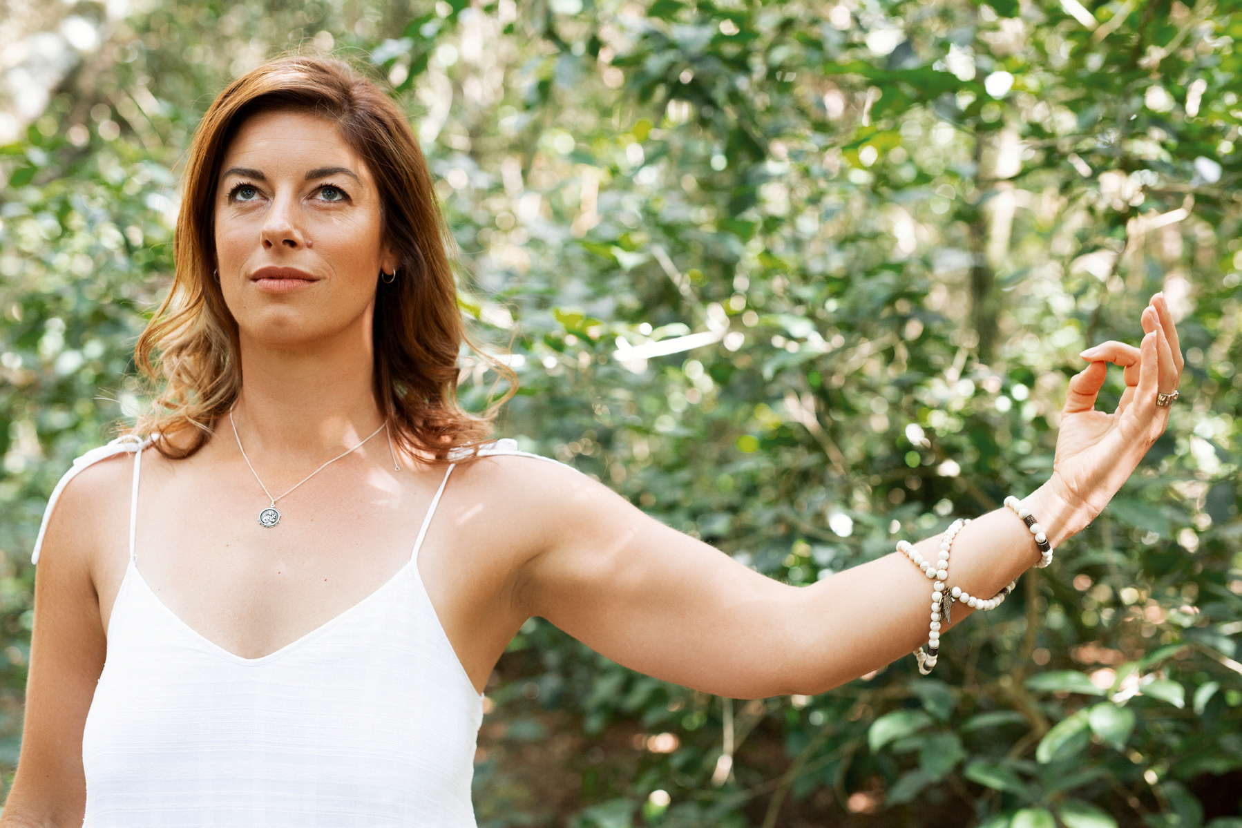 A slim woman stands in front of greenery, one arm outstretched with her index finger and thumb touching 