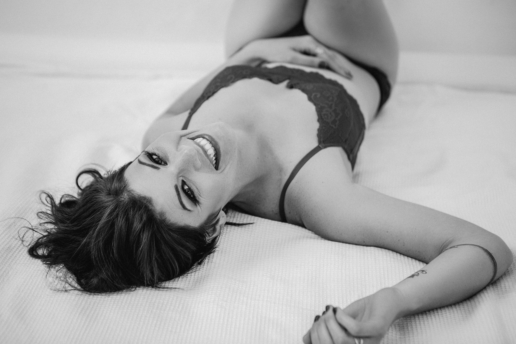 A stunning brunette woman reclines on a bed boudoir shoot style, smiling widely at the camera above her