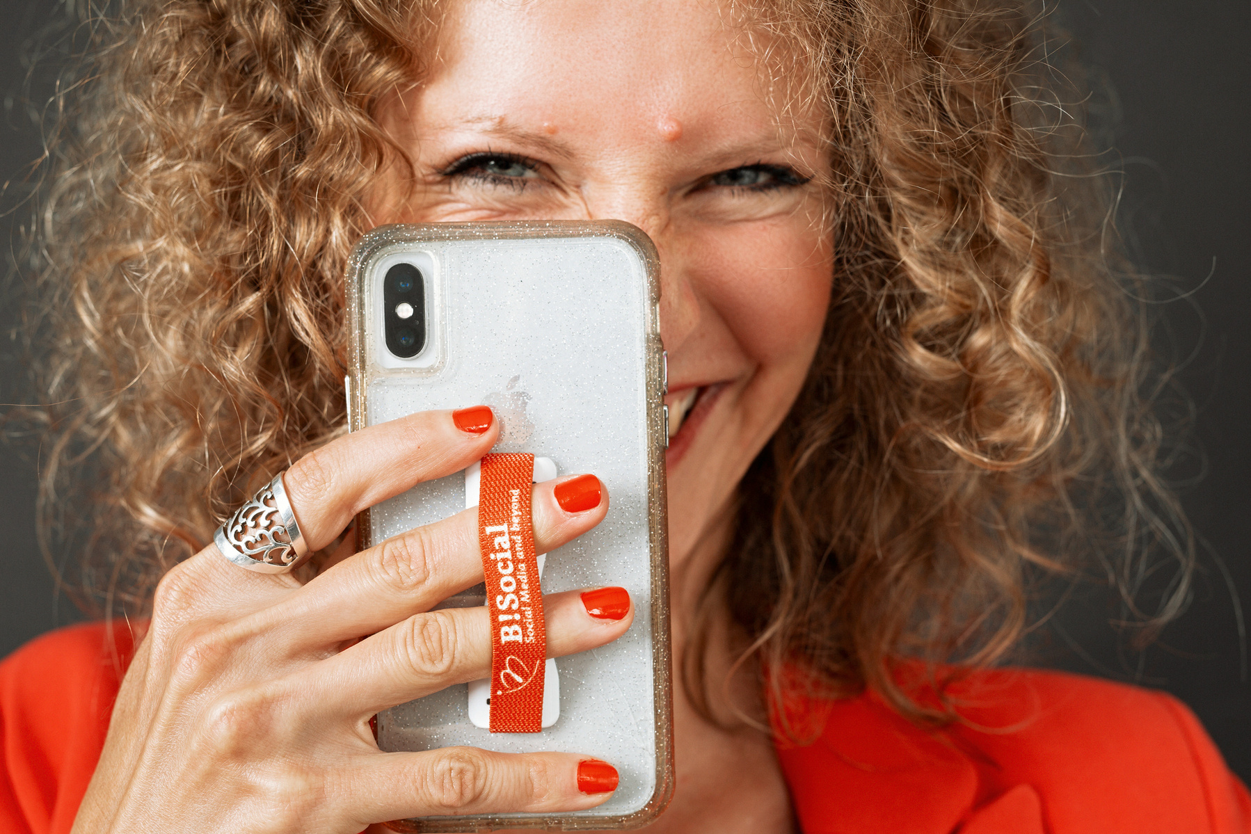 A woman with blonde curls smiles from behind her phone, it looks as though she is snapping a photo of you