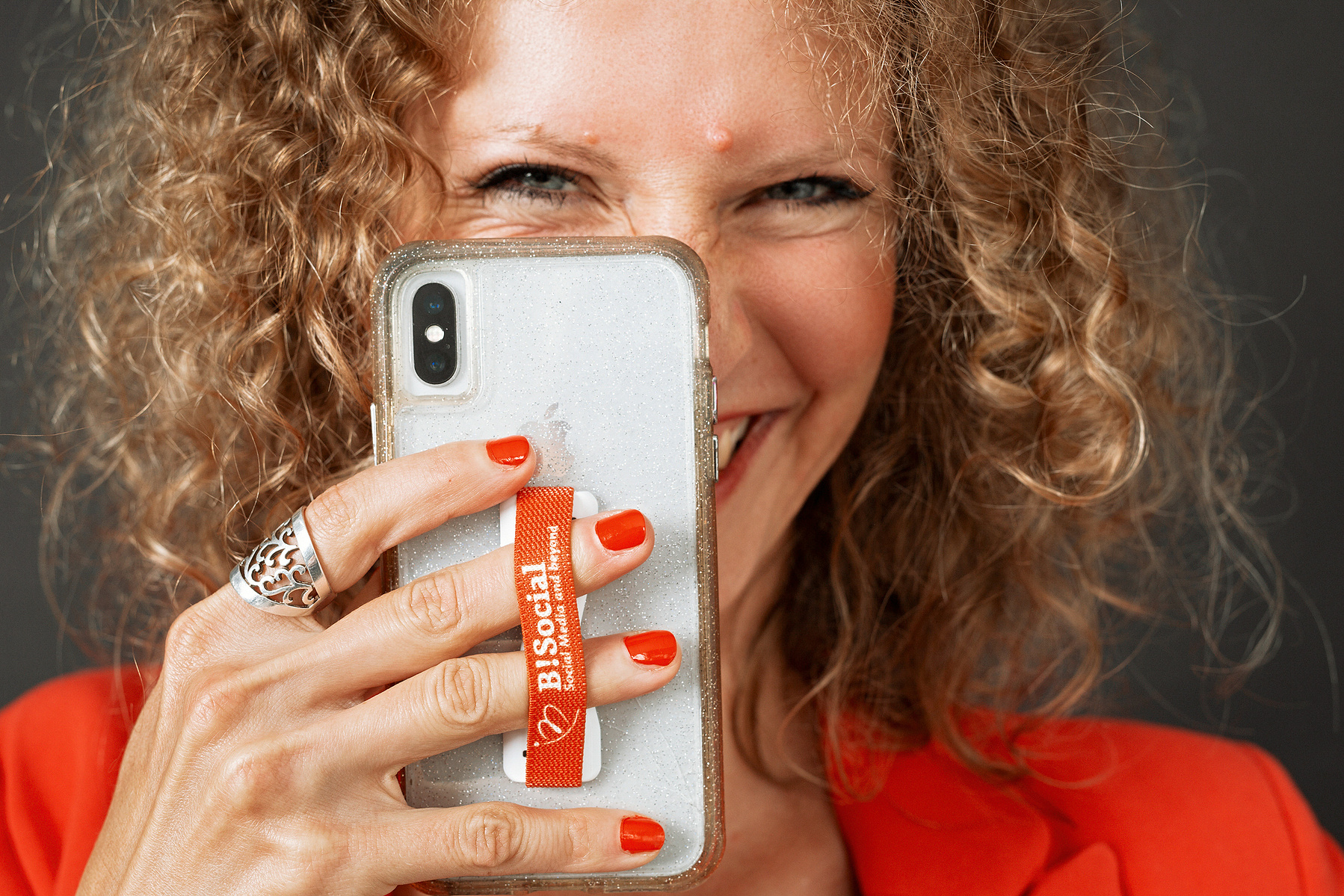 A woman with blonde curls smiles from behind her phone, it looks as though she is snapping a photo of you