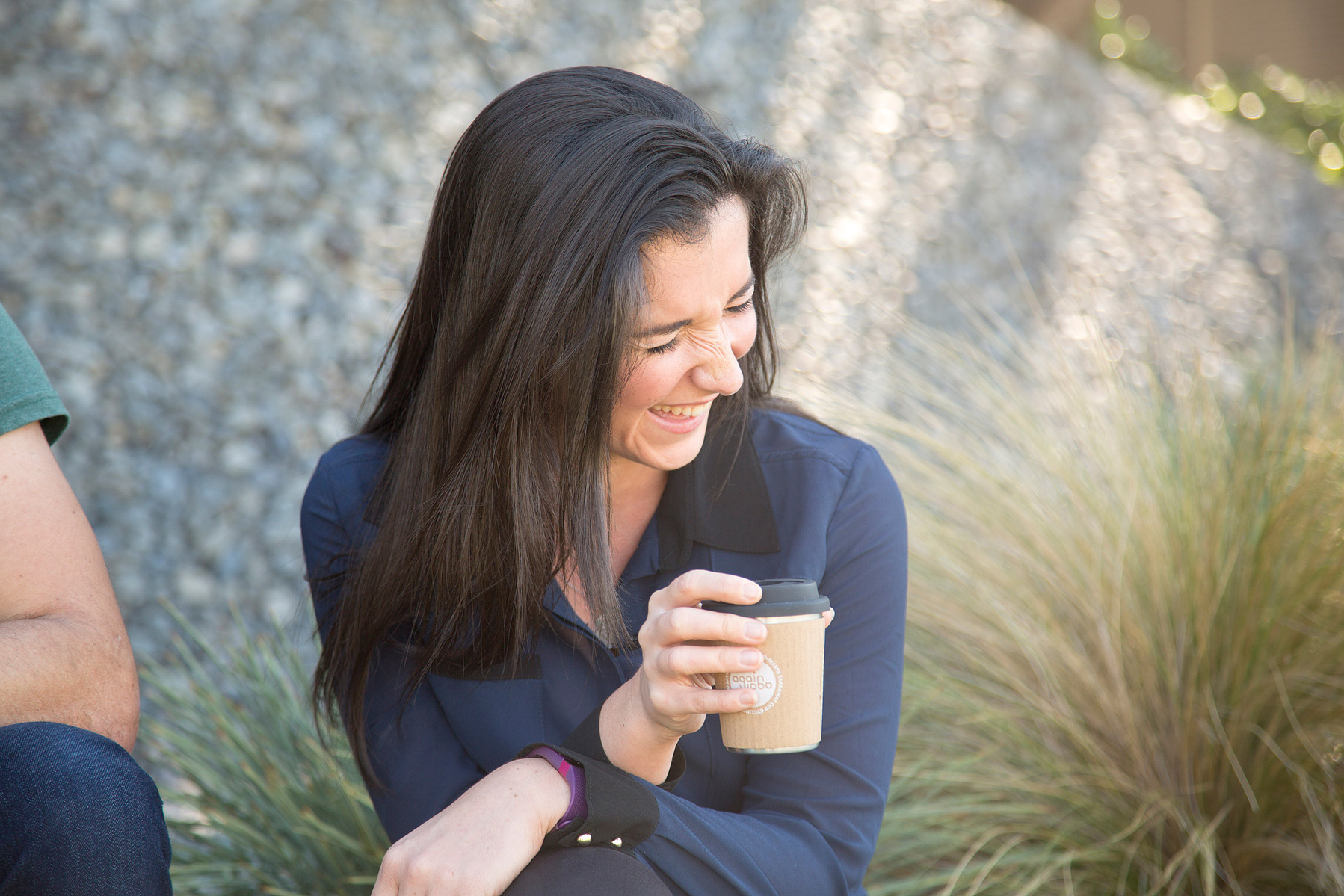 A brunette woman sits and holds a coffee in a takeaway cup, she is laughing at something