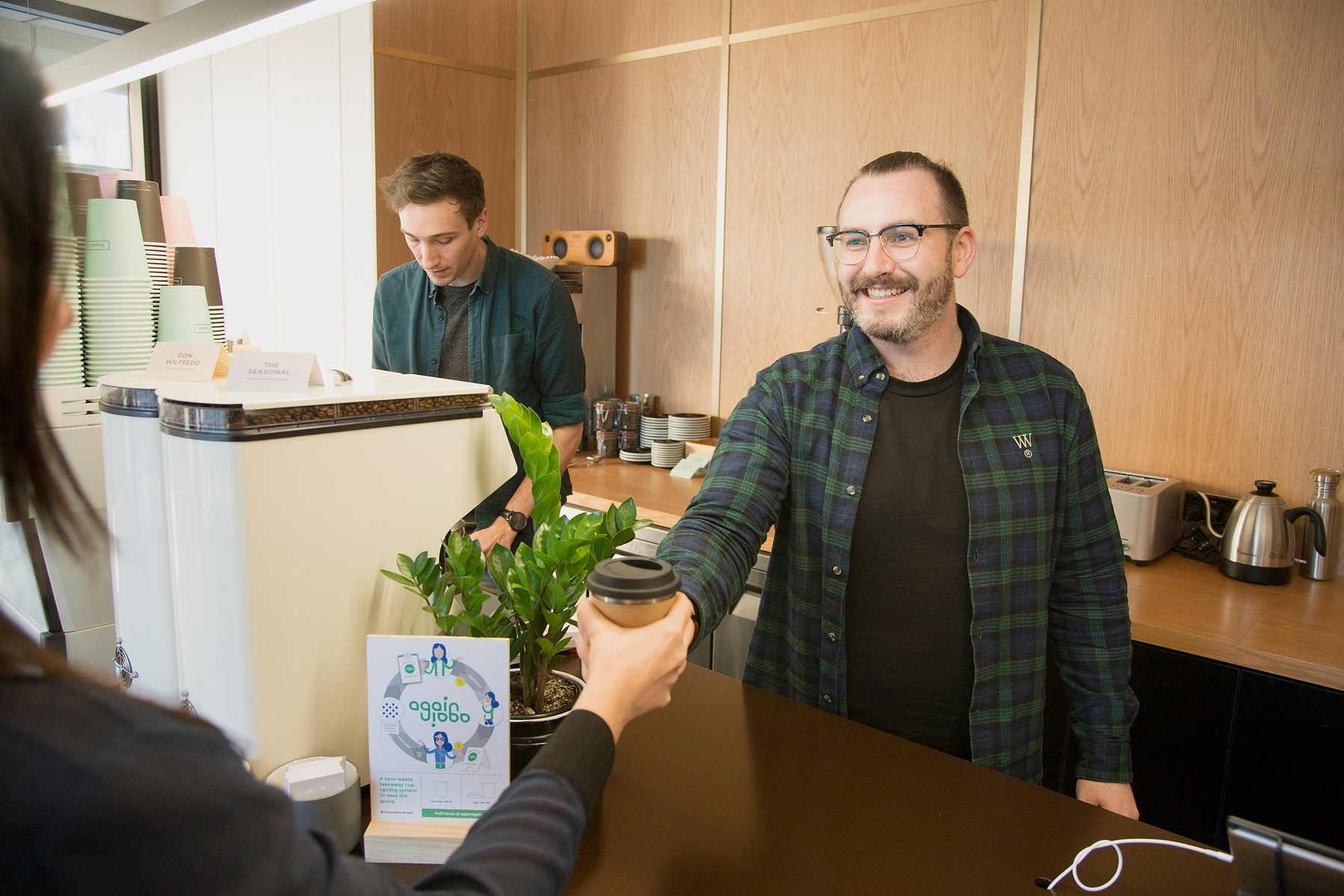 Two people work in a coffee shop, one is making coffee and the other is serving customers with a smile
