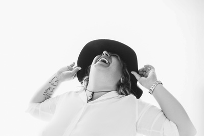A curvaceous woman wearing a white shirt and a wide brimmed hat throws her head back in laughter