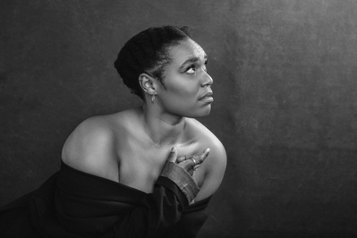An elegant young woman wears a dress of the shoulder, slightly crouched as she looks up towards the right corner of the photo light illuminating her face and upper torso