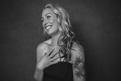 A tattooed blonde woman touches her chest as she smiles off camera
