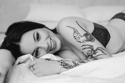 A tattooed young brunette woman lays on a bed, smiling coyly