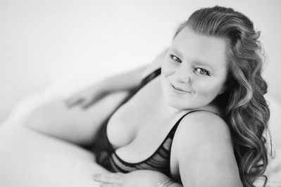 A curvaceous brunette woman reclines onto a bed while smiling at the camera