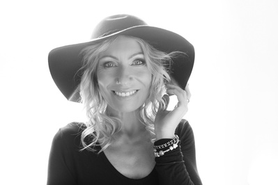 A blonde woman wears a wide brimmed hat, smiling down at the camera with a bright backlight illuminating her