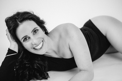 A slim brunette woman wears a bodysuit and reclines on a bed with chin resting on one hand