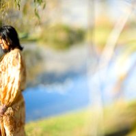 A brunette woman stands by a lake, she looks off camera and the lake is reflecting the mountains behind her