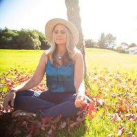 A blonde woman sits outside meditating, the bring sunshine filling the frame