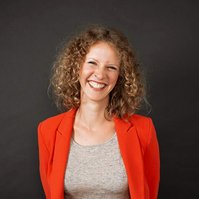 A slim woman with curly hair wears an orange blazer and smiles broadly into the camera