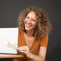 A woman wearing an orange top sits behind a wooden table with a laptiop on, she is holding a sign that spells 'hello' and is smiling