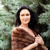 A brunette woman stands in front of frosty pines, she is wrapped in a fur jacket
