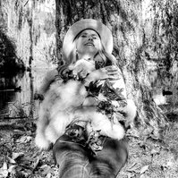 A blonde woman smiles at the sky, she wears a hat and is illuminated in dappled sunlight