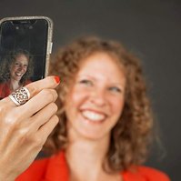 A happy woman with strawberry blonde curls takes a selfie, only the screen of her phone is in focus so we can see the photo she is taking