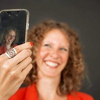 A happy woman with strawberry blonde curls takes a selfie, only the screen of her phone is in focus so we can see the photo she is taking