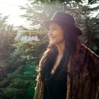 A brunette woman wears a fur jacket and black hat, she is outside and the sun cascades through trees