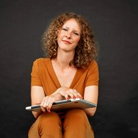 A slim woman sits with her laptop on her knees, a half smile on her lips as she looks at the camera