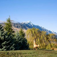 A wide shot of a brunette woman in a floral dress, behind her are the mountains ranges of Queenstown